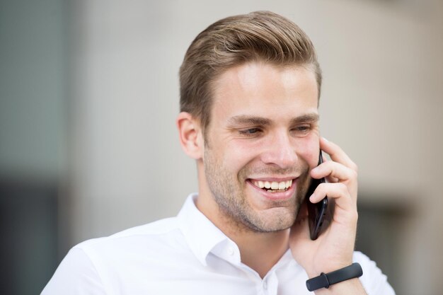 Sempre disponível. Camisa branca bem preparada de homem fala fundo urbano de telefone. Trabalhador de escritório atraente bonito cara fala telefone celular. Empresário sério feliz chamada móvel durante a hora do almoço.