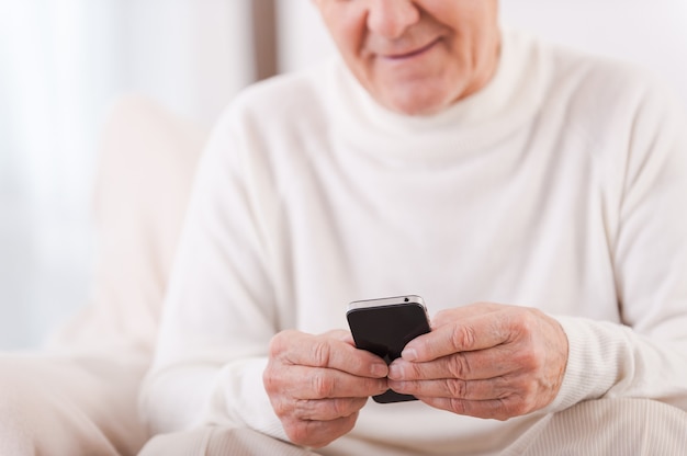 Sempre em contato com o mais próximo. Close de um homem alegre sênior, segurando o celular, enquanto está sentado na cadeira