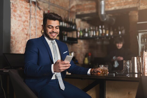 Sempre mantendo contato. Homem de negócios confiante e alegre sentado no balcão do bar, bebendo uísque e fazendo networking no smartphone, copie o espaço