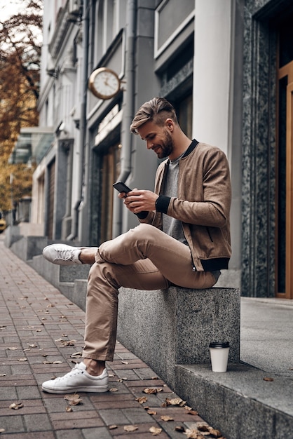 Sempre online. Jovem bonito em uso casual usando seu telefone inteligente enquanto está sentado ao ar livre