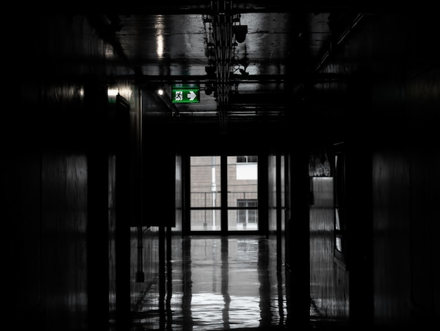 Señal de incendio de salida verde colgada en el techo en un pasillo oscuro y misterioso en el edificio cerca de la puerta de salida de incendios Perspectiva de la habitación de la puerta en un edificio solitario y tranquilo con luz en estilo vertical en blanco y negro