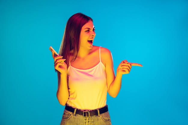 Señalando con el teléfono, sonriendo. Retrato de mujer joven caucásica sobre fondo azul en luz de neón. Modelo de mujer hermosa en casual. Concepto de emociones humanas, expresión facial, ventas, publicidad. Copyspace.