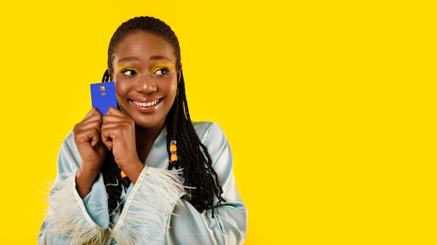 Senhora africana segurando cartão de crédito olhando de lado sobre fundo amarelo