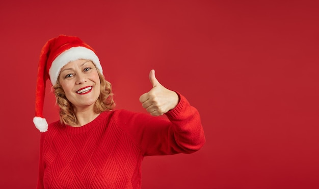 Senhora alegre e sorridente com um chapéu de Papai Noel com uma camisola sobre um fundo vermelho levanta um polegar. Copie o espaço para o seu anúncio