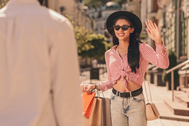 Senhora alegre em dia de sol acenando para um homem e carregando sacolas de compras