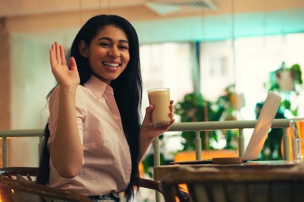 Senhora amigável e positiva sentada com um copo de café com leite em um café e sorrindo enquanto olha para longe e acenando com a mão