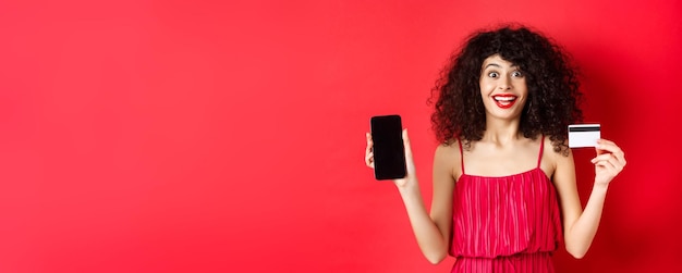 Foto senhora animada em vestido vermelho mostrando cartão de crédito de plástico e tela de smartphone sorrindo espantada em pé