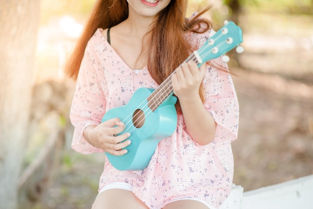 Senhora asiática com chapéu toca música ukulele bossanova no verão Jovem linda mulher tocando música ao ar livre Uma jovem feliz gosta de tocar ukulele debaixo de uma árvore