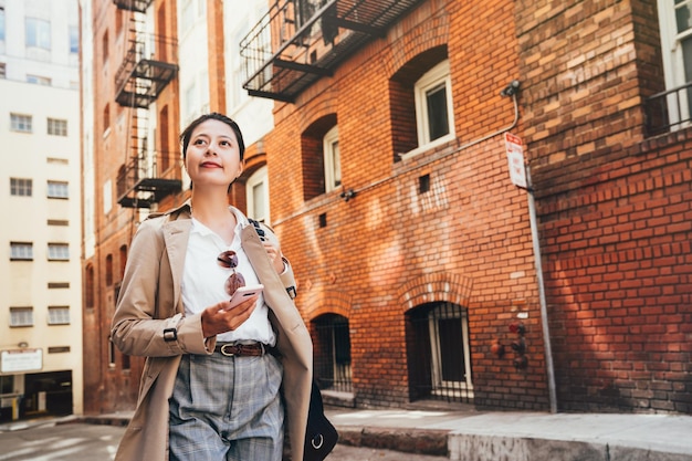 senhora asiática de volta para casa depois do trabalho à tarde. mulher chinesa confiante segurando o celular olhando ao redor vivendo no tradicional apartamento de tijolo vermelho em san francisco. estilo de vida de pessoas urbanas da cidade