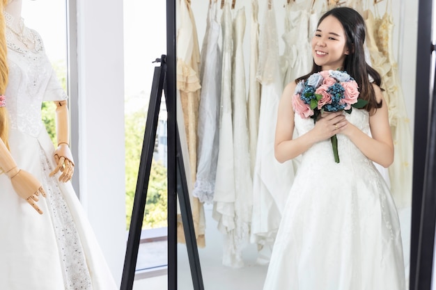 Senhora asiática está olhando no espelho e sorrindo enquanto escolhe vestidos de noiva na loja.