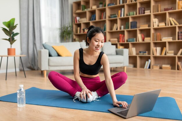 Senhora asiática magra escolhendo tutoriais on-line no laptop para treinar em casa sentado no tapete de ioga em