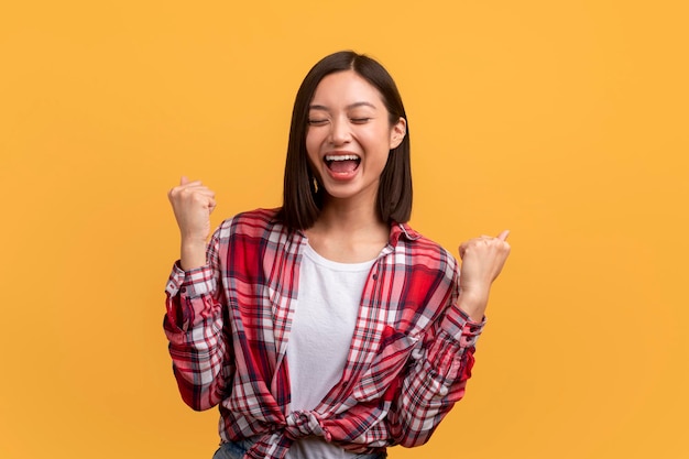 Senhora asiática muito feliz gesticulando sim e apertando os punhos gritando de emoção sobre o fundo amarelo do estúdio