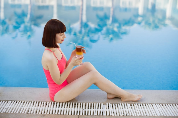 Senhora bonita com bebida alcoólica sentada perto da piscina azul