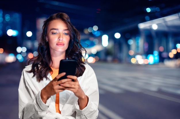 Senhora caucasiana séria enviando mensagens de texto via celular em pé na rua da cidade com iluminação noturna ligada