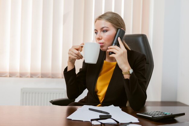 Senhora chefe falando ao telefone e bebe café enquanto está sentado à mesa em seu escritório