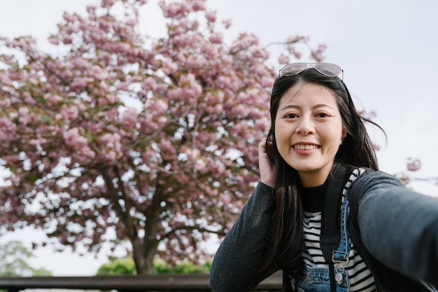 Senhora chinesa de frente para a câmera está sorrindo e pressionando o cabelo dela. turista asiático tomando selfie com linda sakura em plena floração.