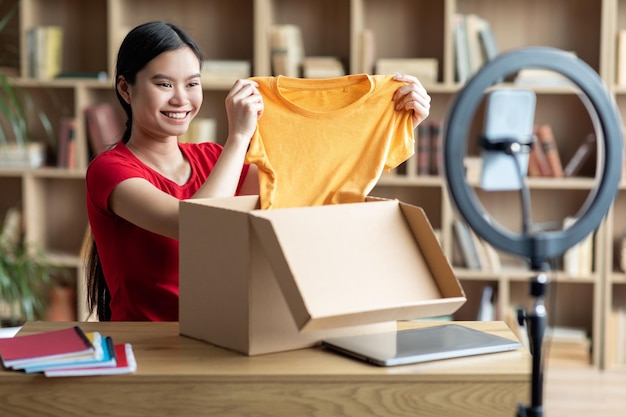 Senhora chinesa milenar sorridente tira roupas da caixa de papelão e olha para a webcam do smartphone