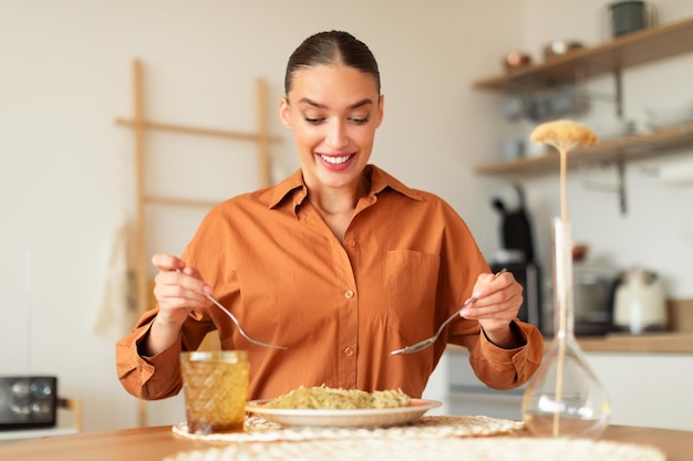 Senhora com fome comendo segurando garfo e colher comendo macarrão italiano caseiro sentado no interior da cozinha