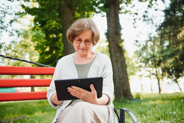 Senhora confiante positiva posando com tablet no parque Mulher de cabelos grisalhos sênior em casual sentado no banco do parque e usando o conceito de conexão sem fio do tablet