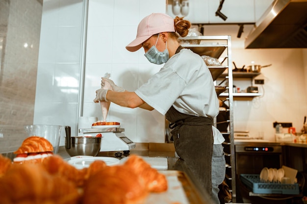 Senhora de máscara aplica creme rosa em croissant na bancada na padaria