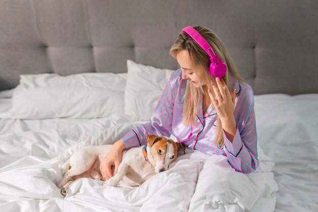 Senhora despreocupada de pijama ouvindo música em fones de ouvido enquanto está sentada na cama com seu amado animal de estimação