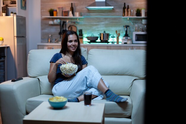 Senhora divertida assistindo programa de tv à noite, sentada no sofá