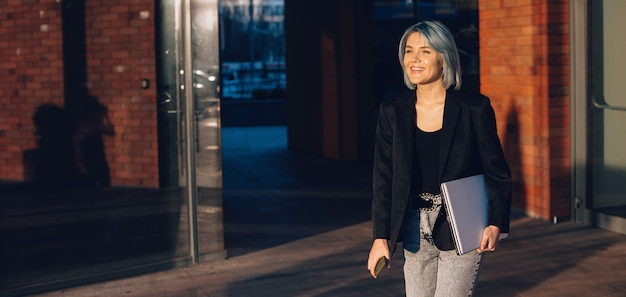 Senhora encantadora andando na rua com um laptop, sorrindo e segurando o telefone