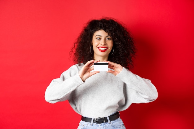 Senhora feliz com cabelo encaracolado, mostrando o cartão de crédito de plástico e sorrindo, de pé contra a parede vermelha. Conceito de compras.