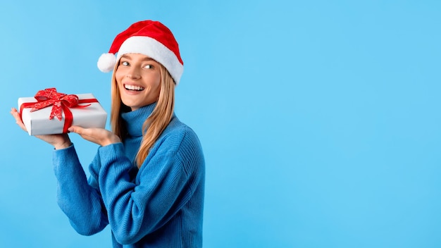 Senhora feliz de suéter e chapéu de Papai Noel segurando uma caixa de presente e olhando para o espaço livre do estúdio azul