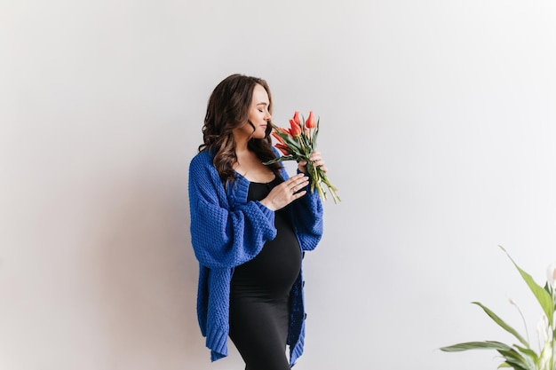 Senhora feliz de vestido preto cheira tulipas Mulher morena grávida de casaco azul segura flores sobre fundo branco