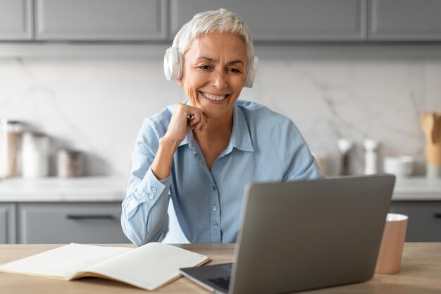 Senhora idosa alegre em fones de ouvido assistindo palestra no laptop interno