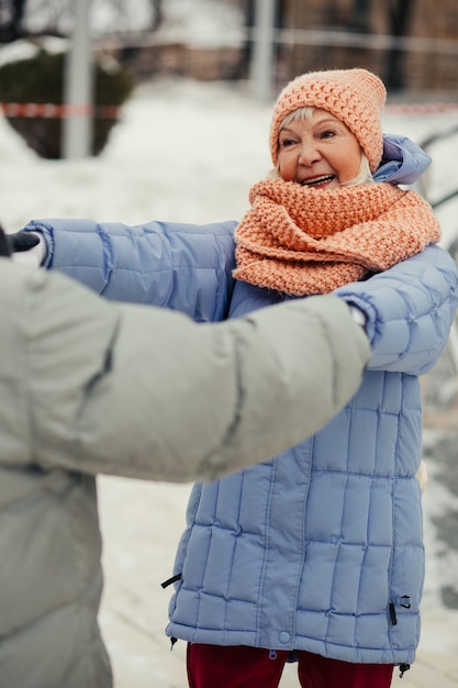 Senhora idosa com casaco de inverno sorrindo para o marido enquanto segura suas mãos e dança com ele na rua