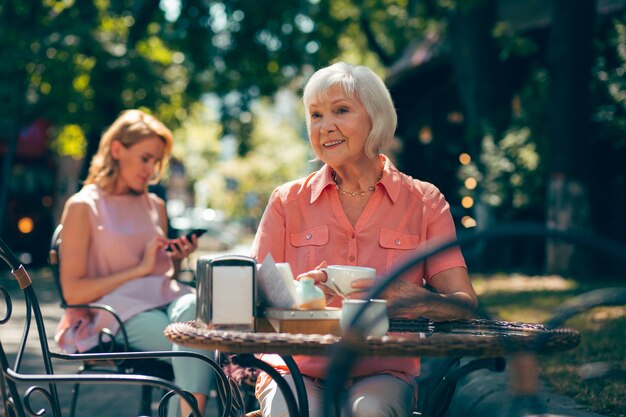 Senhora idosa positiva sorrindo e tomando café em um café de rua