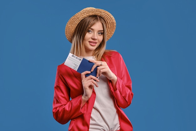 Senhora loira bonita com chapéu de palha, blusa branca e terninho vermelho. Ela está sorrindo, segurando seu passaporte e passagem