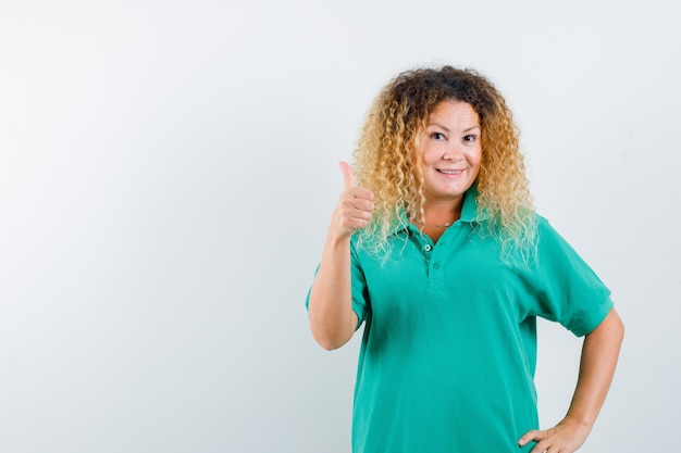 Senhora loira e bonita aparecendo o polegar em uma camiseta polo verde e parecendo feliz. vista frontal.