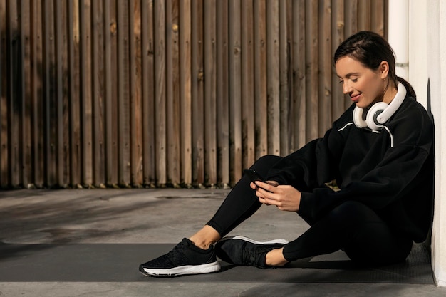 Senhora milenar caucasiana sorridente em roupas esportivas e fones de ouvido descansando sentado no chão digitando no smartphone