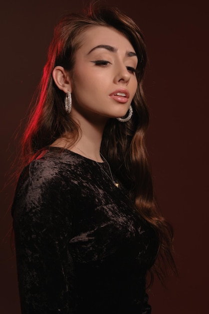 Senhora morena atraente em joias elegantes e vestido de veludo preto posando contra background colorido