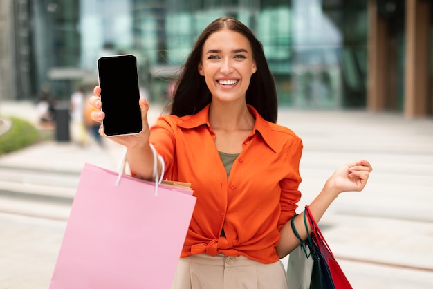 Senhora mostrando a tela do telefone segurando sacolas de compras em área urbana