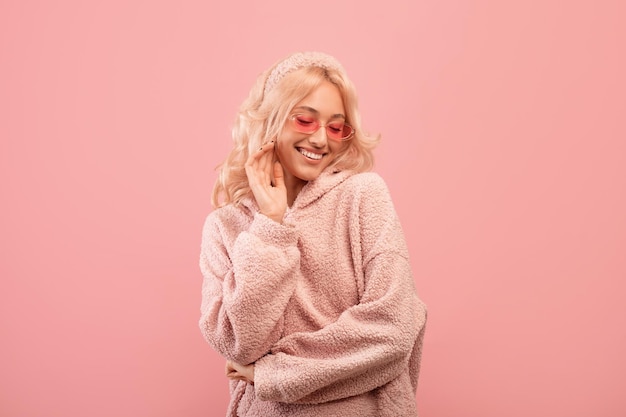 Senhora muito elegante olhando para baixo e sorrindo usando óculos escuros coloridos e capuz posando sobre rosa