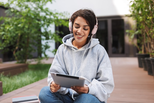 Senhora muito sorridente sentada com fones de ouvido e tablet nas mãos enquanto passa o tempo feliz no pátio da universidade