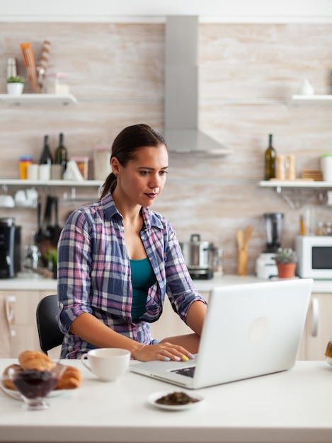 Senhora navegando na internet usando laptop na cozinha e tomando uma xícara de chá verde quente durante o café da manhã