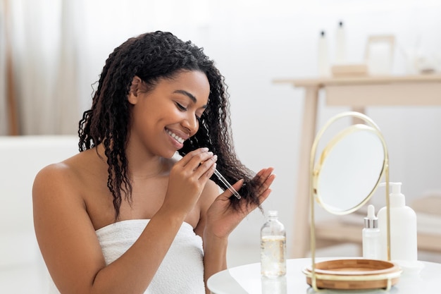 Senhora negra feliz aplicando óleo hidratante nas pontas do cabelo danificado após o banho