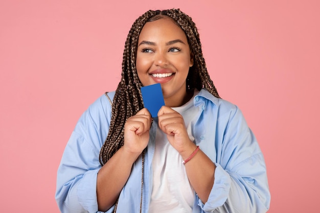 Senhora negra segurando cartão de crédito sonhando com compras fundo rosa