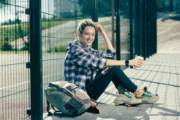 Senhora positiva com dreadlocks segurando seu café e sorrindo enquanto está sentada ao lado da cerca de arame