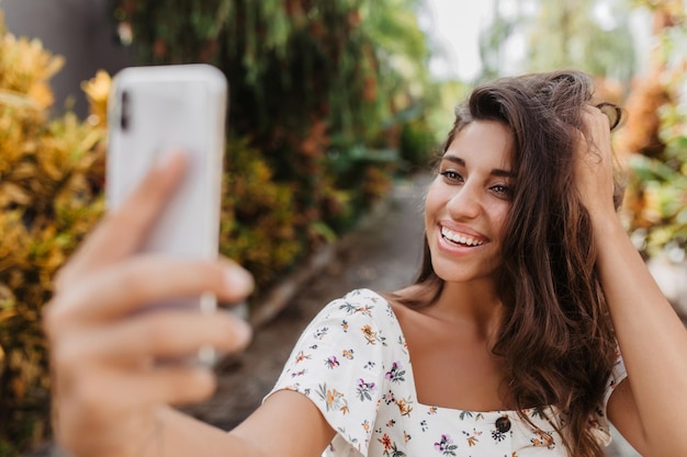 Senhora positiva no topo floral branco segura smartphone e sorridente faz selfie no parque
