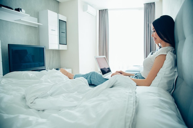 Senhora positiva sentada na cama com um laptop no colo e sorrindo enquanto digita em um teclado