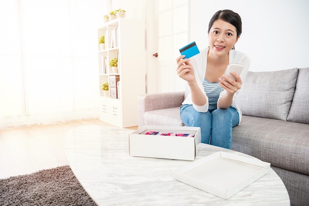 Senhora relaxando no sofá com seu pacote na mesa em sua sala de estar, de frente para a câmera, segurando seu cartão de crédito e celular