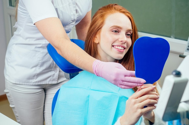 Senhora ruiva ruiva verificando os dentes no espelho no consultório do dentista