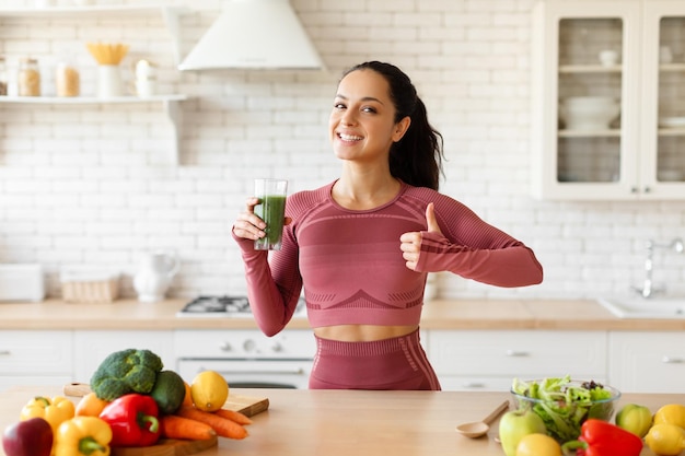 Senhora segurando smoothie verde e gesticulando com o polegar para cima na cozinha