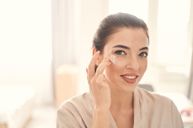 Senhora sorridente e alegre que passa um tempo com procedimentos de beleza em casa pela manhã, enquanto usa um creme suave para os olhos